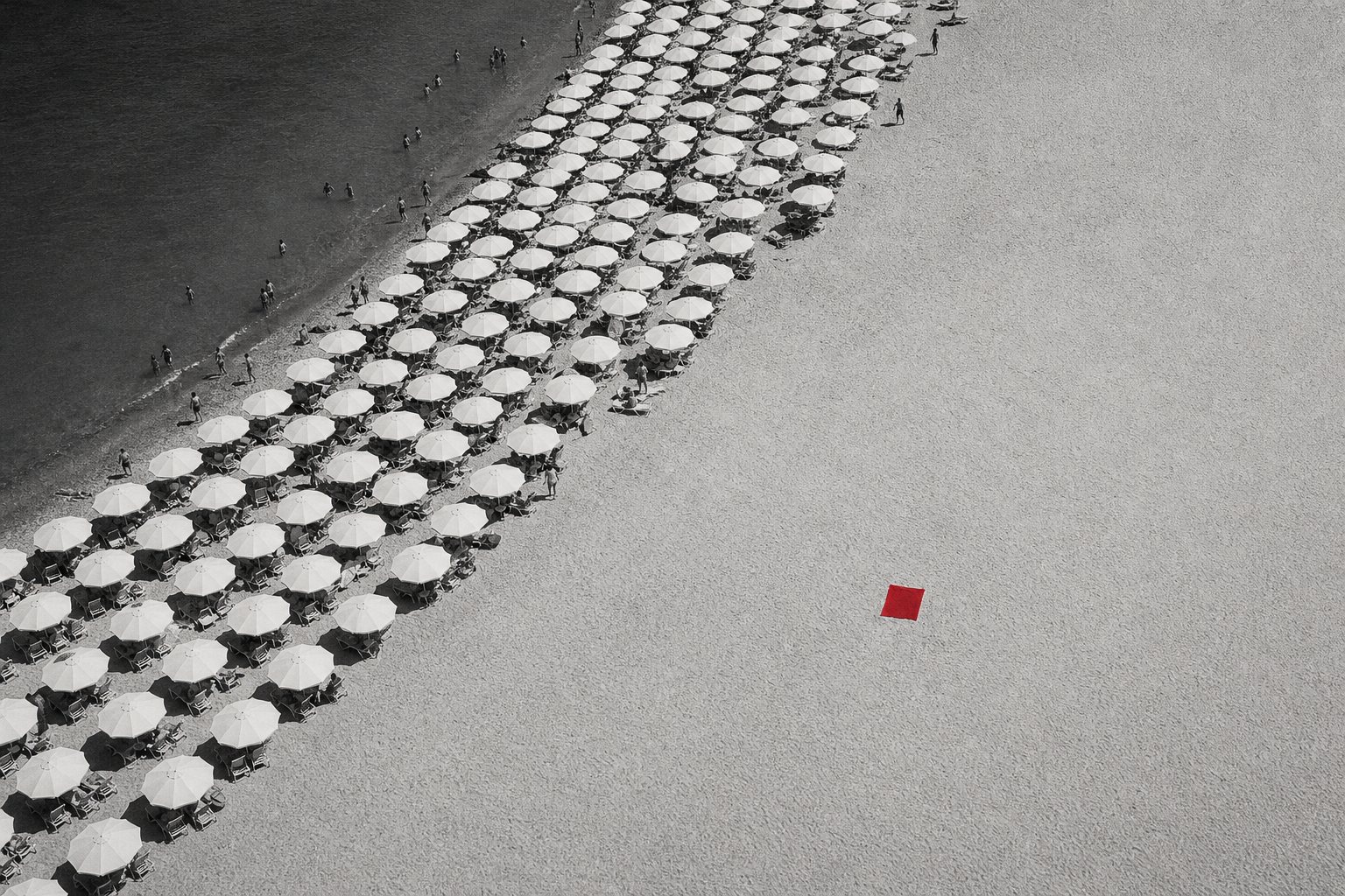 Aerial black-and-white photo of a beach where hundreds of identical white parasols crowd one side while a single red square towel lies alone on the wide empty sand.